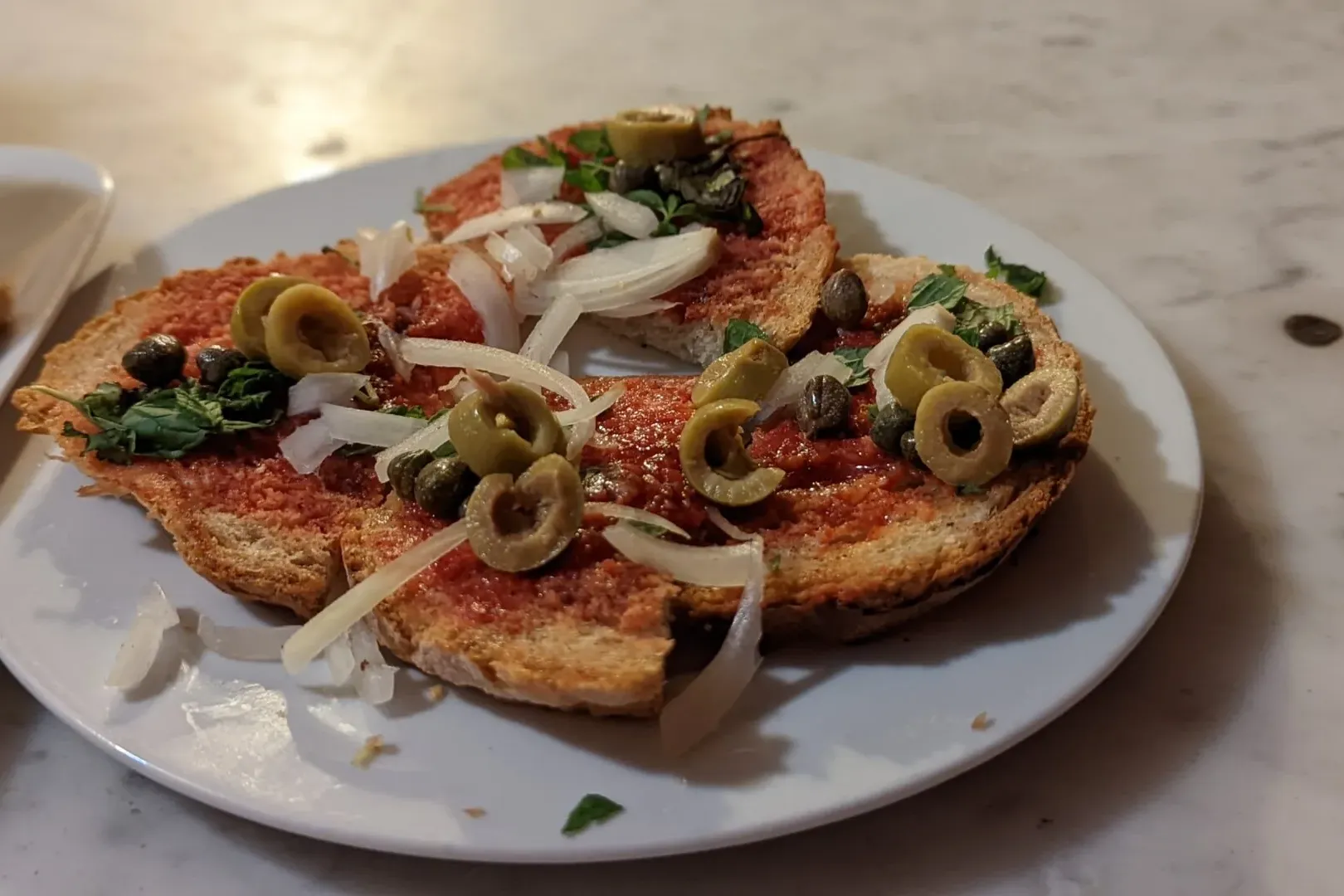 Rebanadas de pan tostado con pasta de tomate rojo, aceitunas verdes, cebolla, alcaparras y hierbas en un plato blanco.