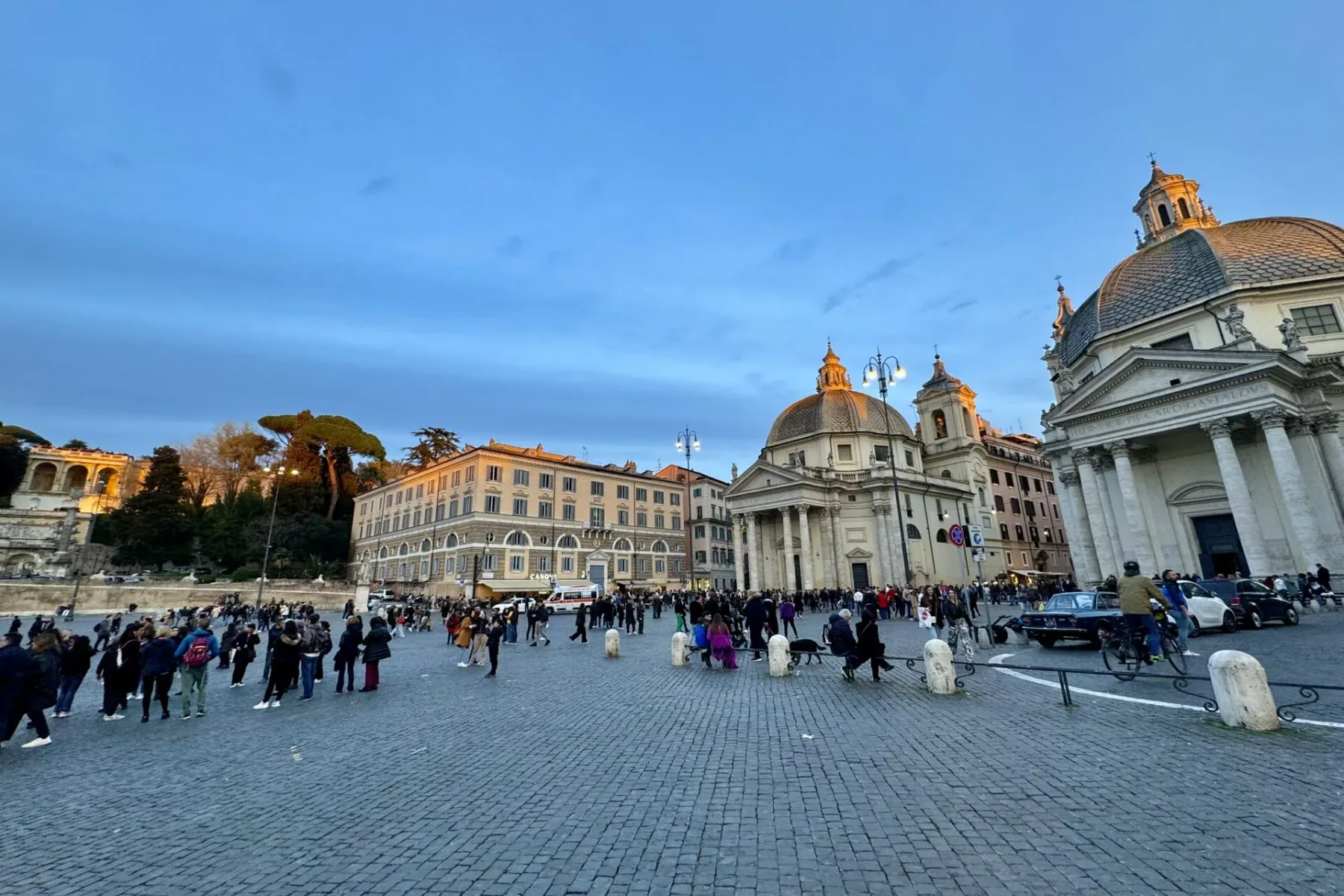 Piazza del Popolo in Rom mit zwei Kuppelkirchen und vielen Menschen auf dem Kopfsteinpflasterplatz bei Dämmerung.