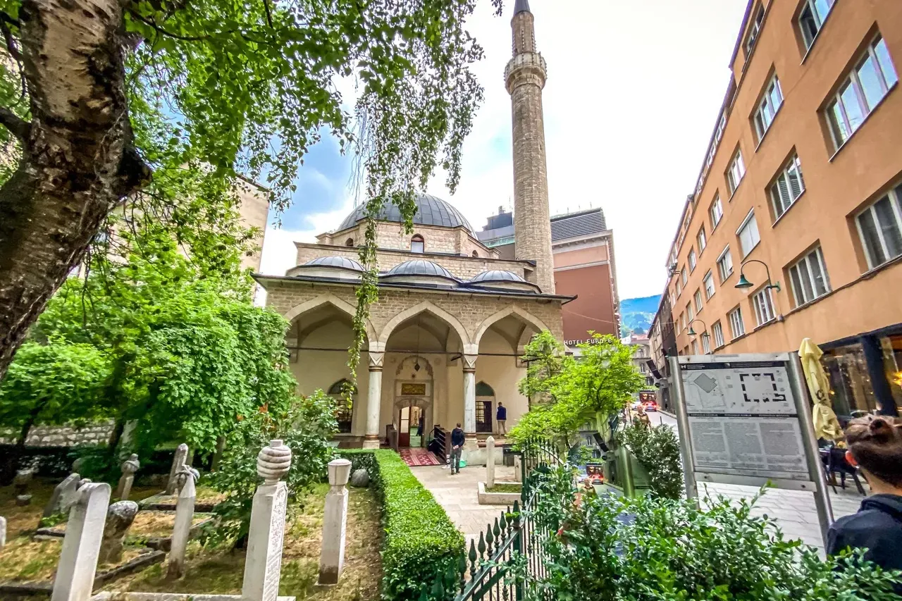 Historic Gazi Husrev-beg Mosque with a tall minaret and dome, surrounded by a cemetery, green trees, and city buildings.