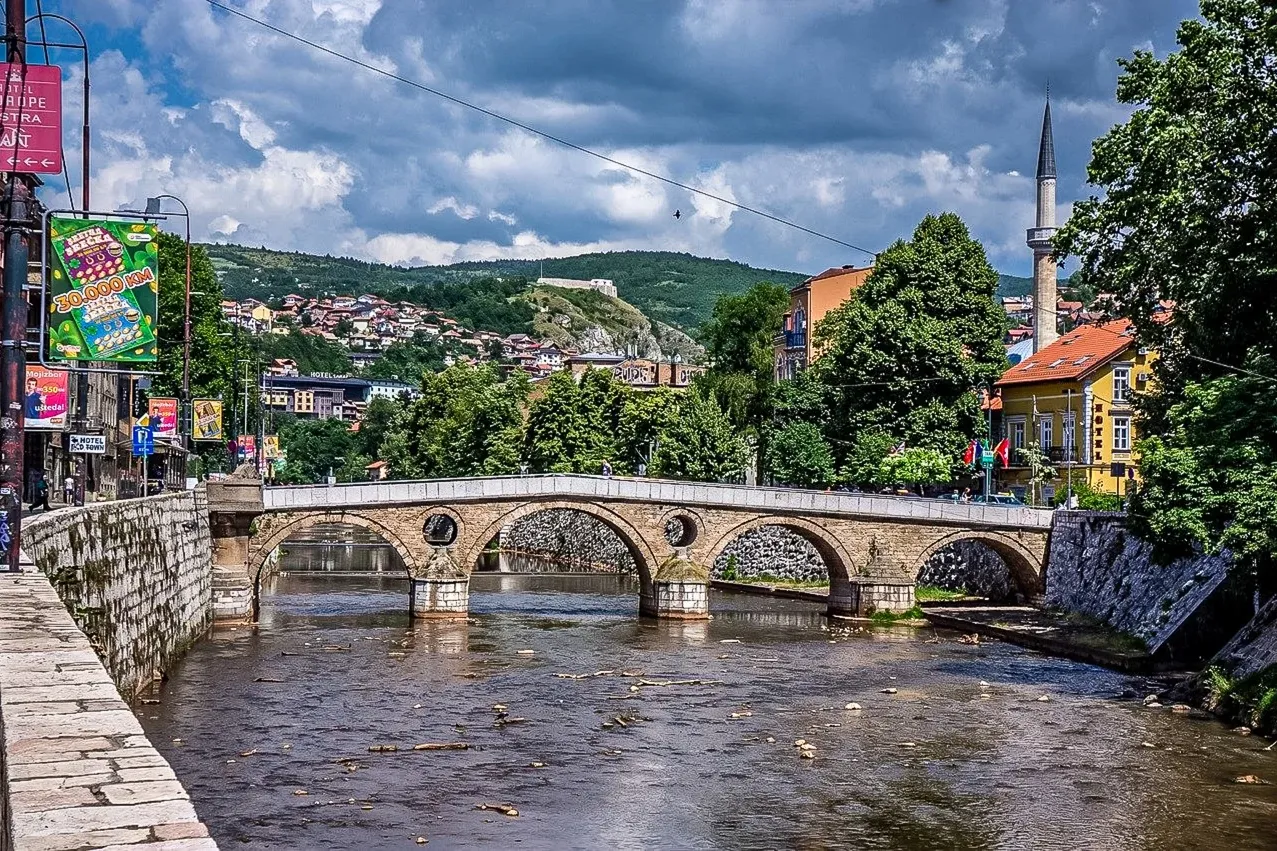 Historic stone arch bridge over a river in Sarajevo, with a minaret, buildings, and green hills under a cloudy sky.