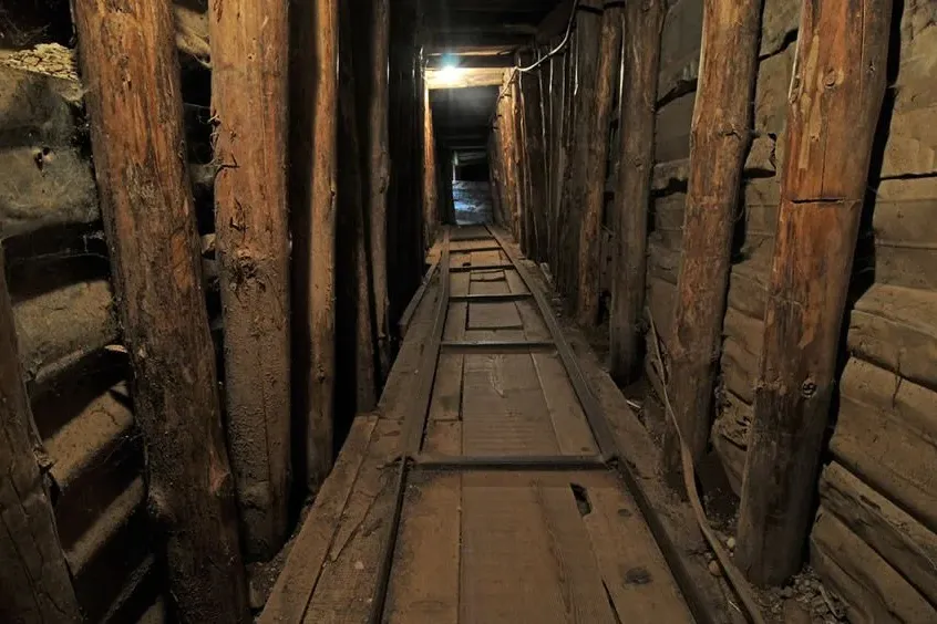 Narrow, dimly lit tunnel with wooden log supports along the walls and a wooden walkway on the floor, extending into the di...