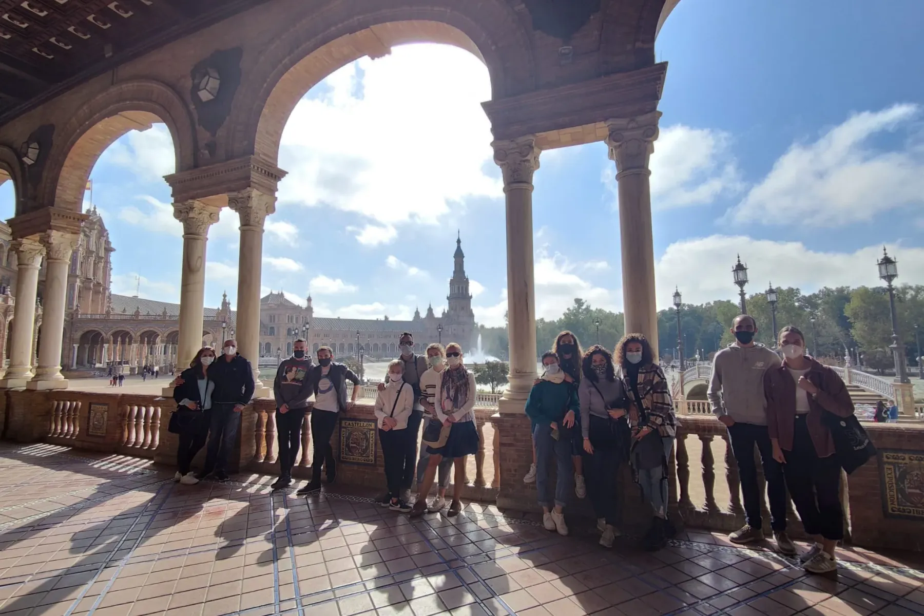 Grupa ludzi, wielu w maskach, stoi pod arkadową kolumnadą, z widokiem na Plaza de España i jej wieżę w Sewilli.