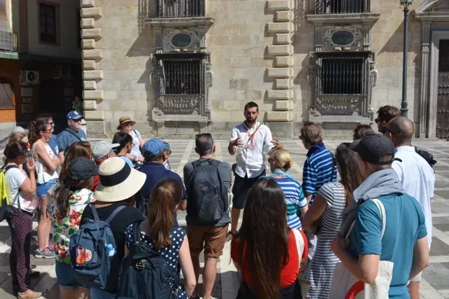 A male tour guide speaks to a group of diverse tourists in front of an ornate stone building with barred windows in Granada.
