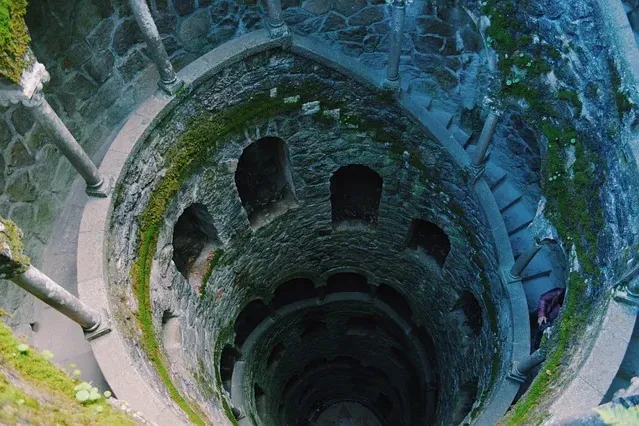 Blick von oben in einen tiefen, moosbewachsenen Steinbrunnen mit einer Wendeltreppe. Eine Person steigt die Stufen hinab.