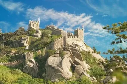 Alte Steinmauern einer Burg und Türme auf einem steilen, grünen, felsigen Hügel unter einem strahlend blauen Himmel mit ve...