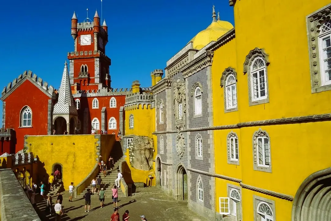 Außenansicht des Pena-Palastes in Sintra mit roten und gelben Abschnitten, einem Uhrturm und Touristen im Hof.