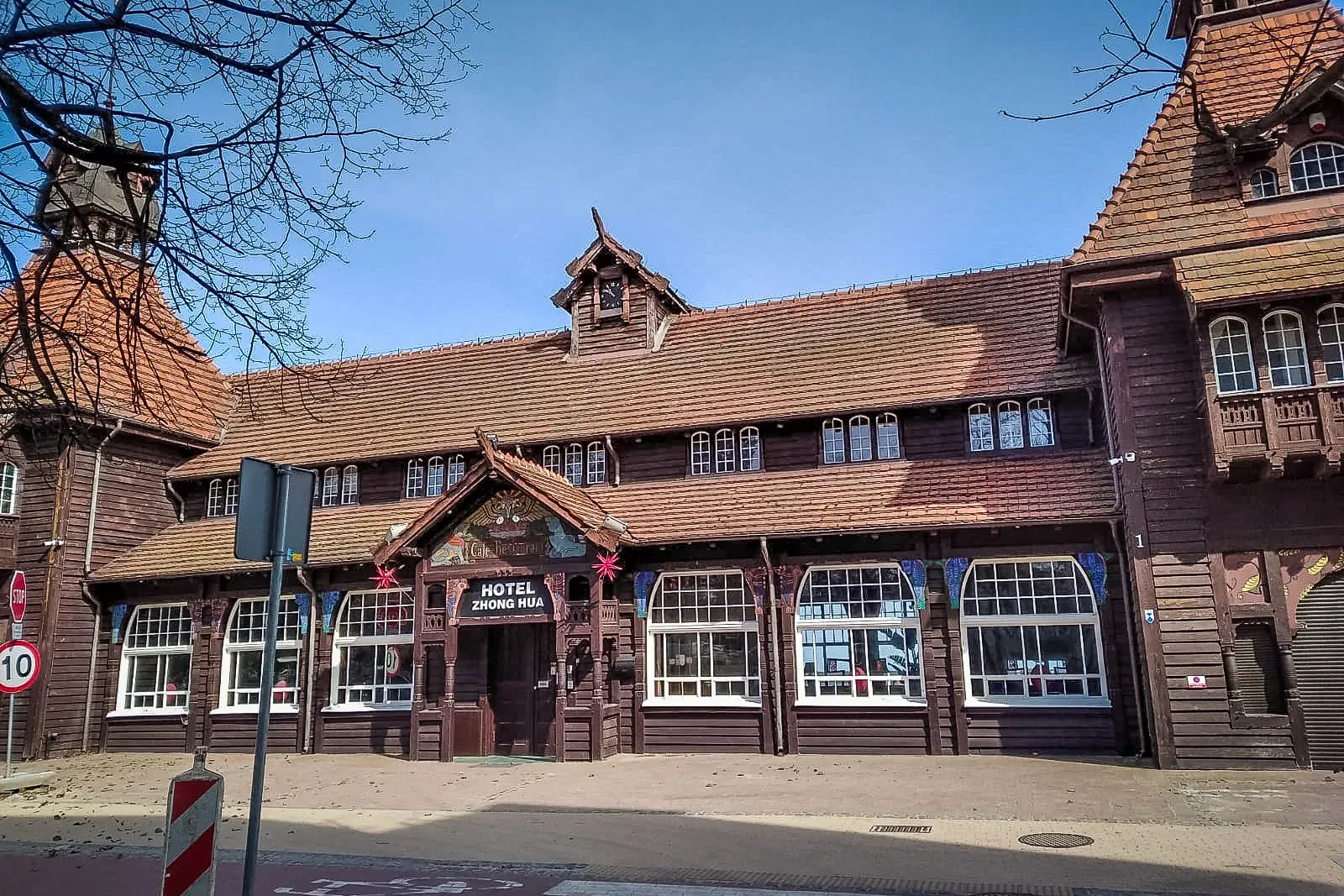Large dark wooden hotel building with brown tiled roof, many white windows, and 'Hotel Zhong Hua' sign. Street and bare br...