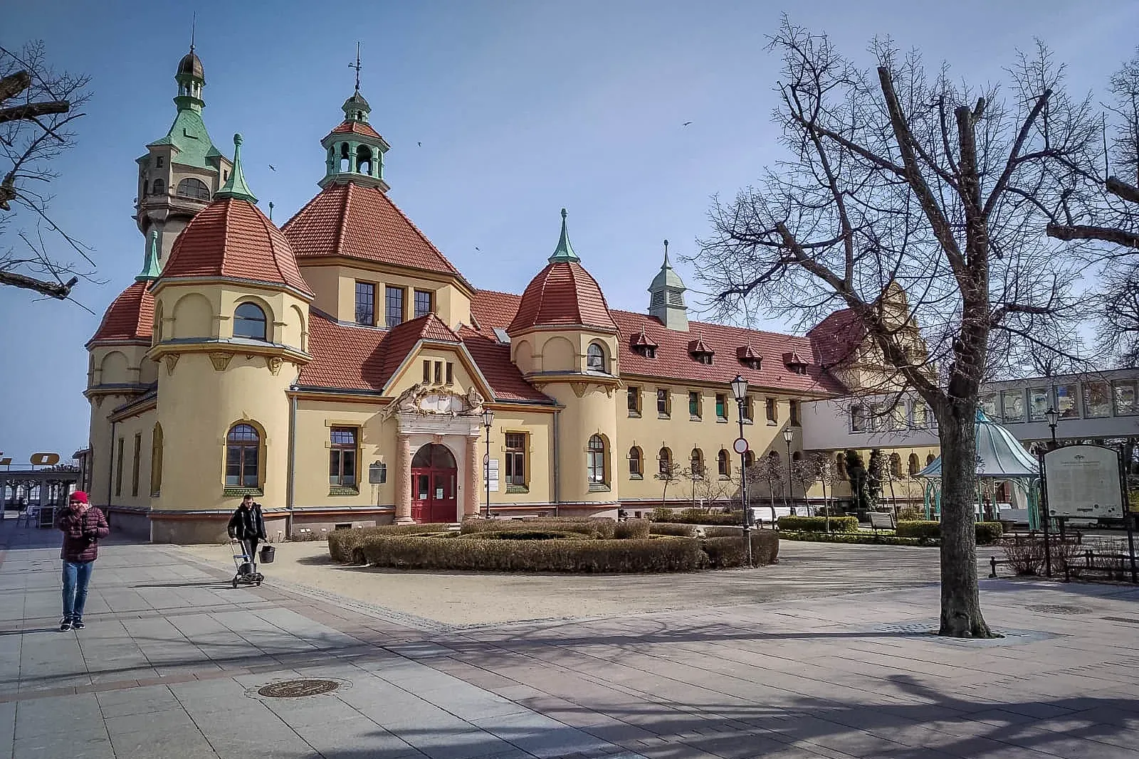 Large cream-colored building with red tiled roofs and green turrets, situated in a paved square with bare trees. Two peopl...