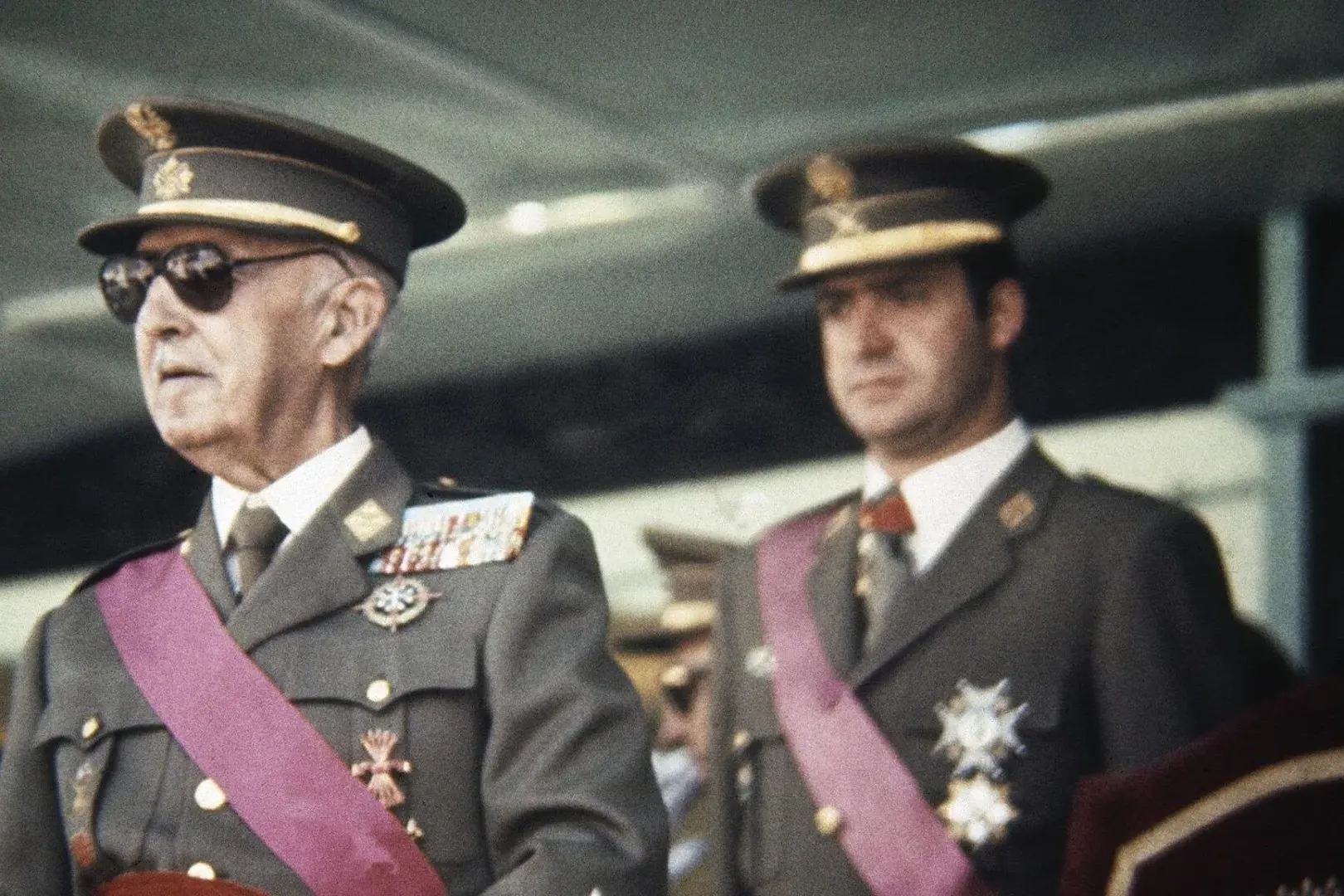Francisco Franco mit Sonnenbrille und Militäruniform mit Orden und violetter Schärpe, dahinter Juan Carlos I.