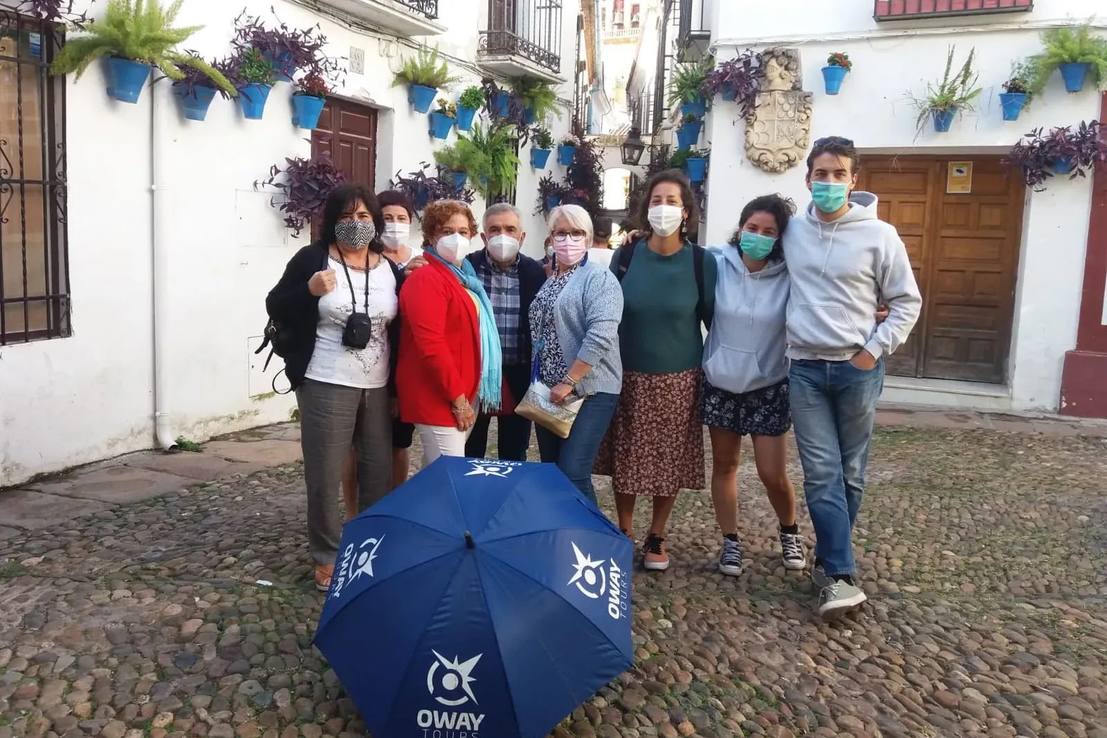 Group of eight people wearing masks standing on a cobblestone street lined with white buildings adorned with blue flower p...