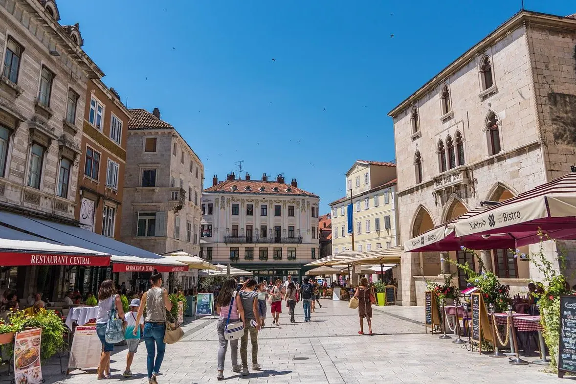 Plaza concurrida del casco antiguo de Split con edificios de piedra, cafeterías al aire libre y gente paseando bajo un cie...