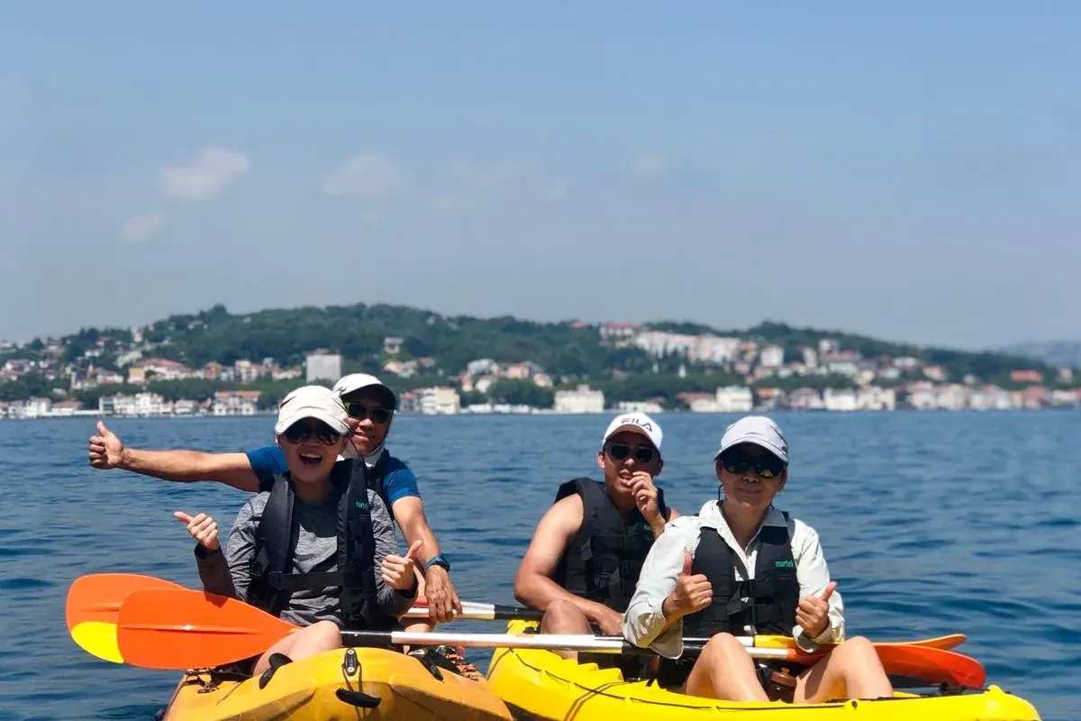 Cuatro personas en dos kayaks amarillos en agua azul, sonriendo y mostrando los pulgares hacia arriba. Una ciudad en la or...
