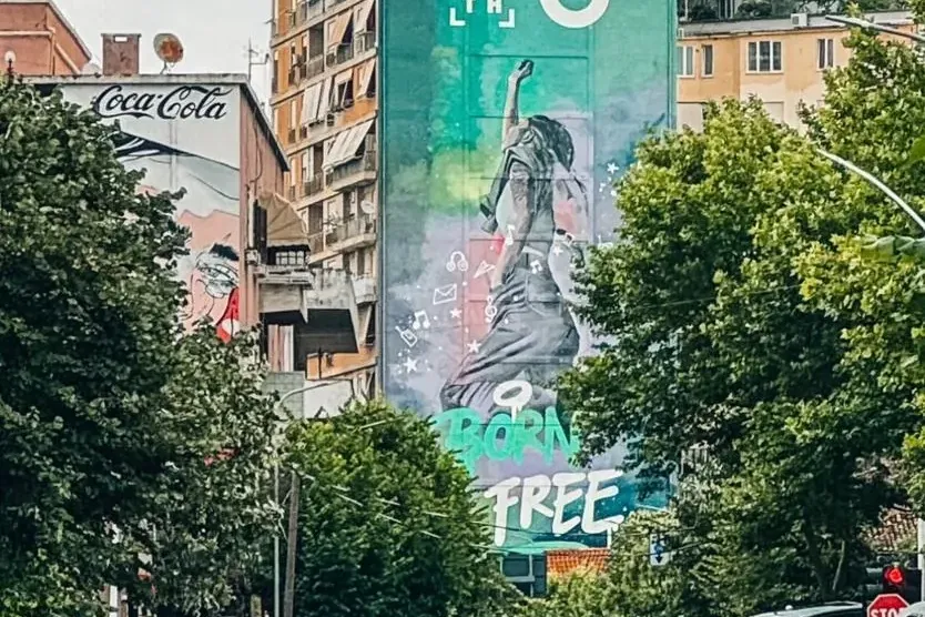 Widok ulicy z samochodami i drzewami; wysoki budynek z dużym muralem postaci i napisem 'BORN FREE', na szczycie kopuła z n...