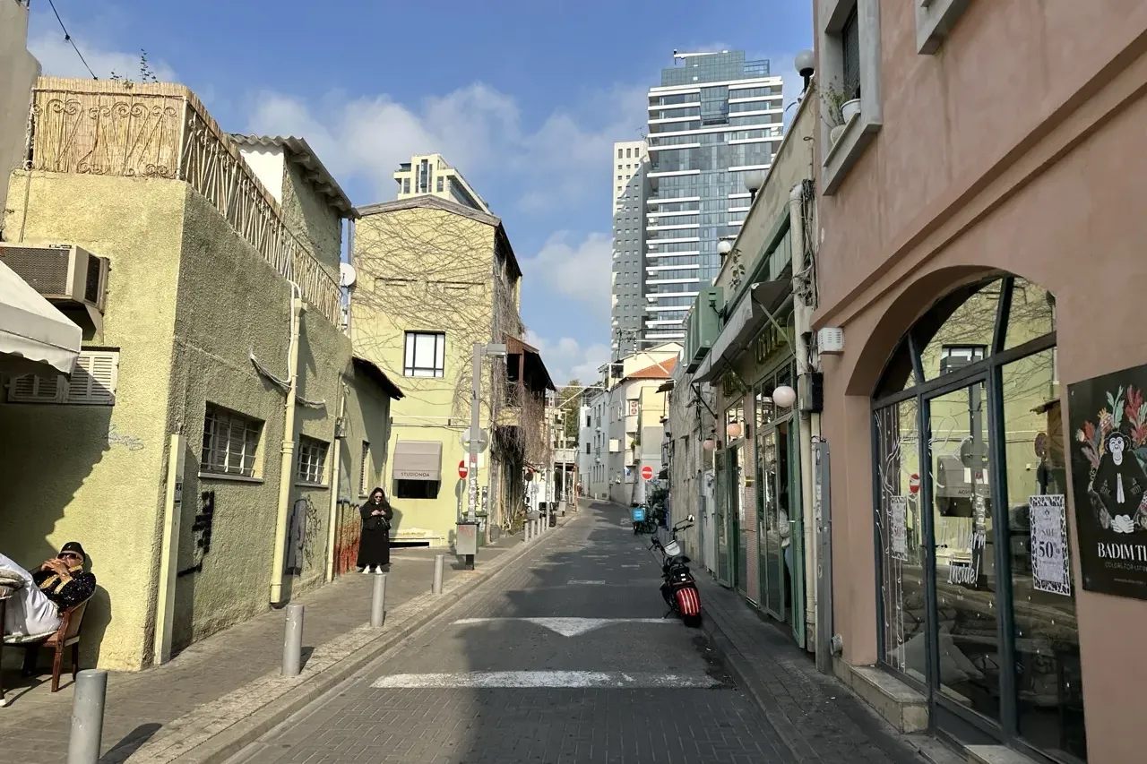Narrow street in Tel Aviv with old, pale buildings and a modern skyscraper in the background under a blue sky.