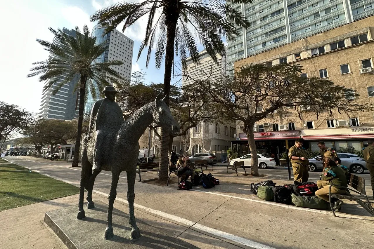 Bronze statue of a man on a horse in Tel Aviv, with palm trees, modern and older buildings, and people in the background.
