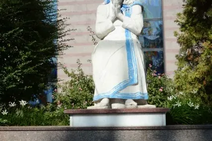 Weiße Statue von Mutter Teresa mit blauen Streifen, gefalteten Händen, auf einem Sockel mit Marmorplatte auf Albanisch.