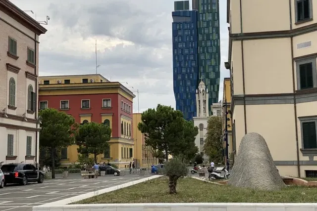 Widok ulicy w Tiranie z nowoczesnym niebiesko-zielonym wieżowcem, tradycyjnymi budynkami, wieżą kościoła i szarą rzeźbą.