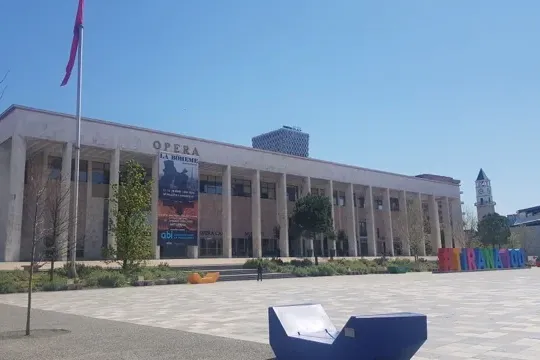Großes neoklassizistisches Gebäude mit 'OPERA LA BOHEME' Banner, ein Platz mit modernen Bänken, Glockenturm und Minarett i...
