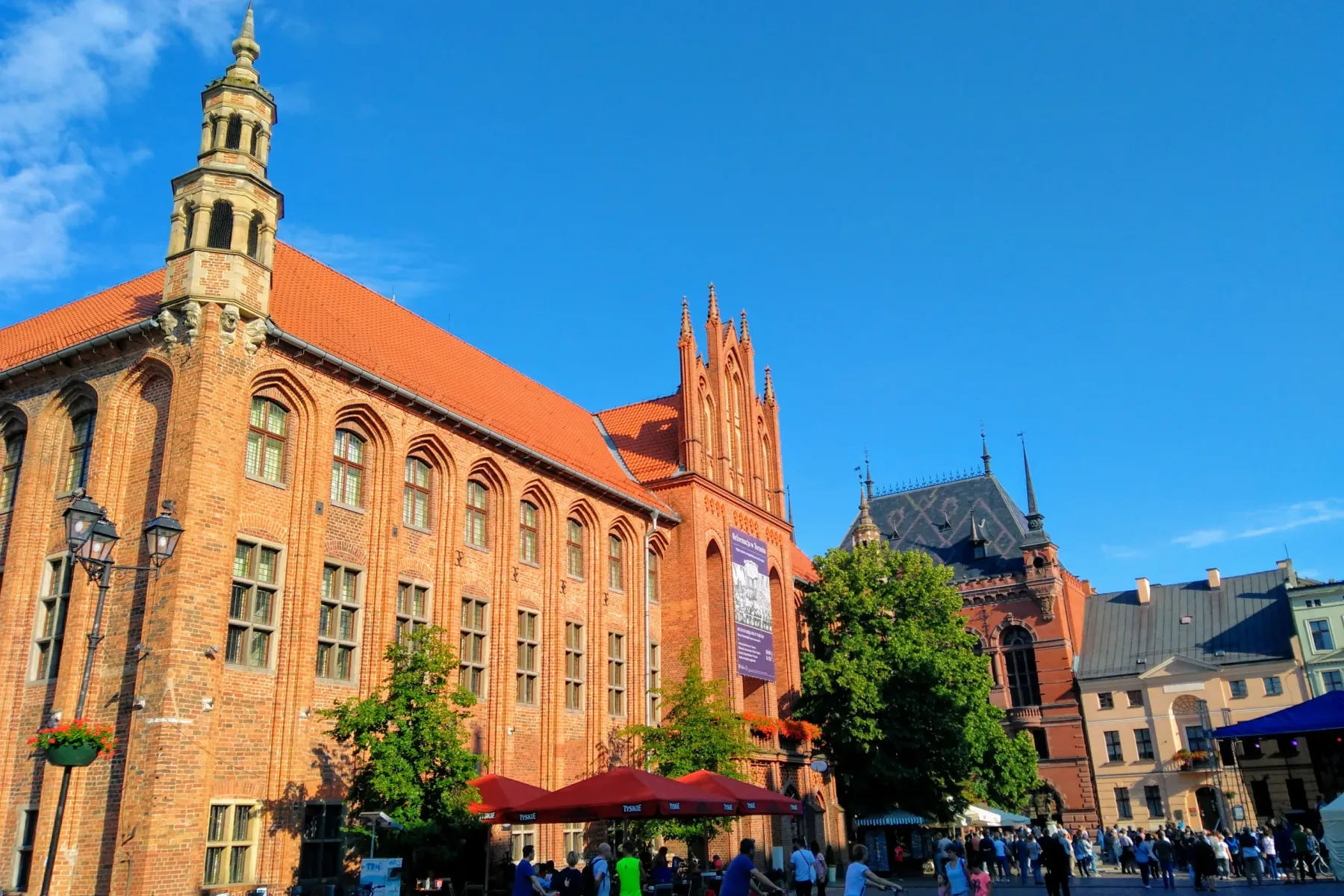 Das Altstädtische Rathaus in Toruń, ein Backsteingebäude mit hohem Turm, auf einem belebten Marktplatz mit Menschen.