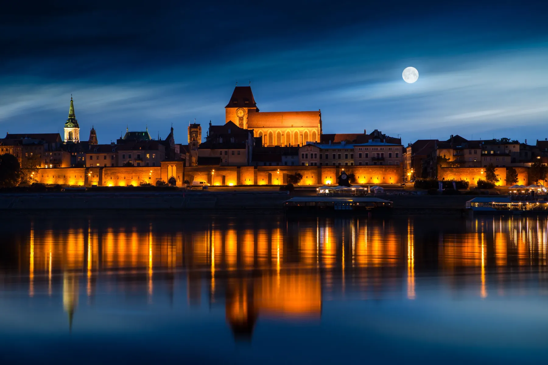 Skyline der Altstadt von Toruń bei Nacht beleuchtet, im Fluss Weichsel gespiegelt unter blauem Himmel mit Vollmond.