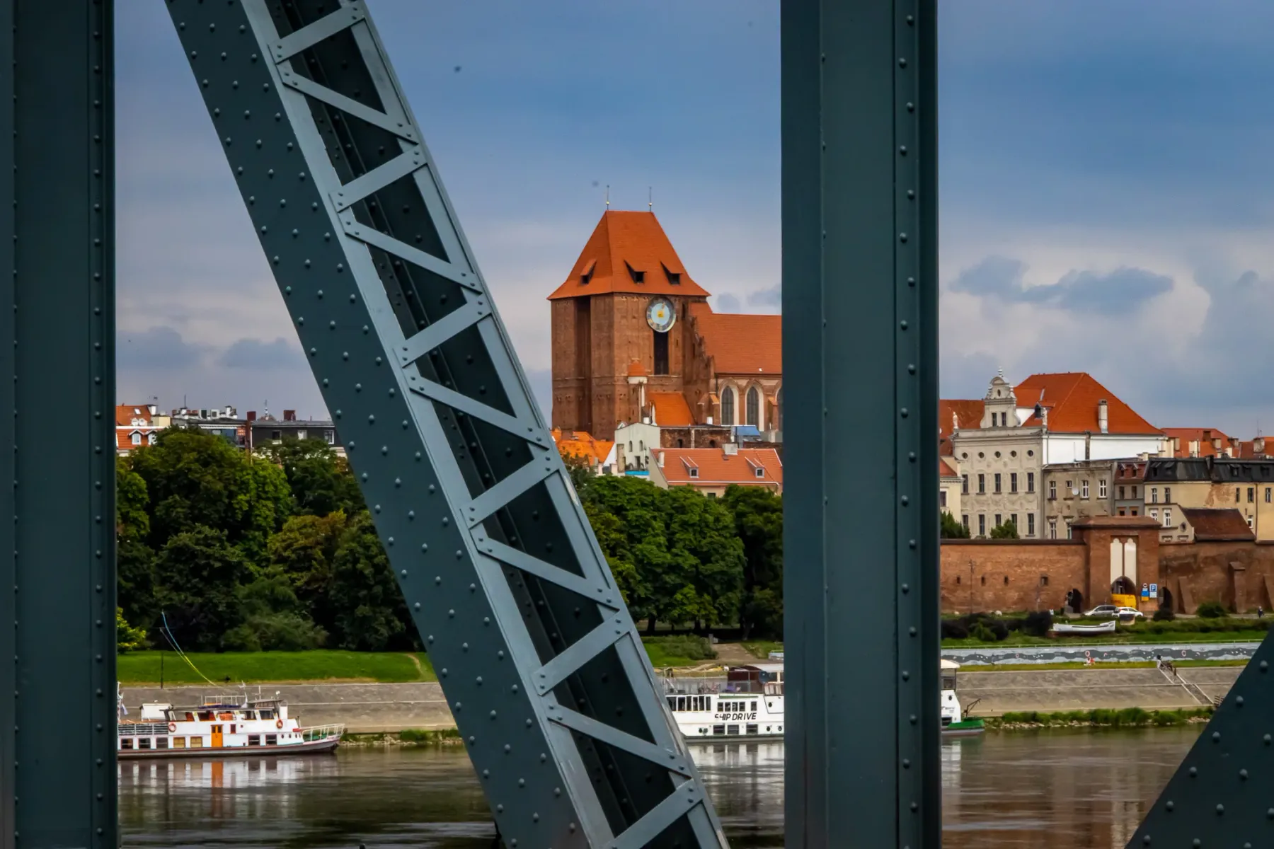 Blick durch eine Brückenkonstruktion auf Toruńs Altstadt mit der Johanneskathedrale und der Weichsel mit Schiffen.