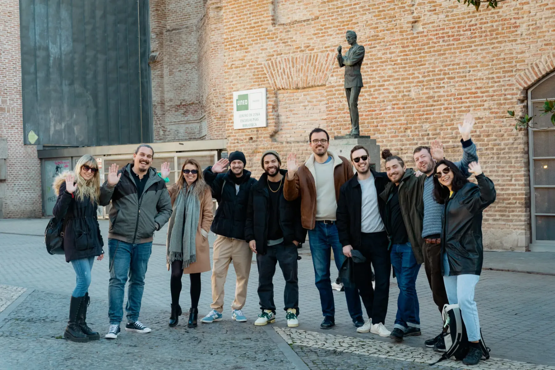 A diverse group of eleven people waving and smiling at the camera on a paved street in Madrid. Behind them is a brick buil...