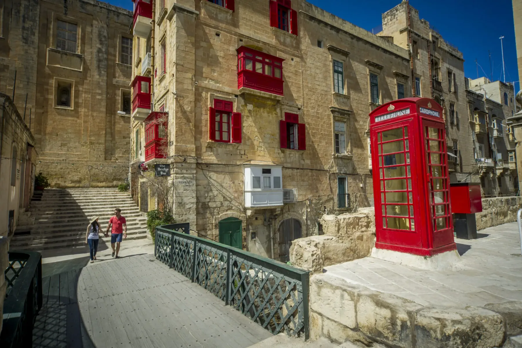 Una pareja camina por un puente en La Valeta, pasando edificios con balcones rojos y una cabina telefónica roja.