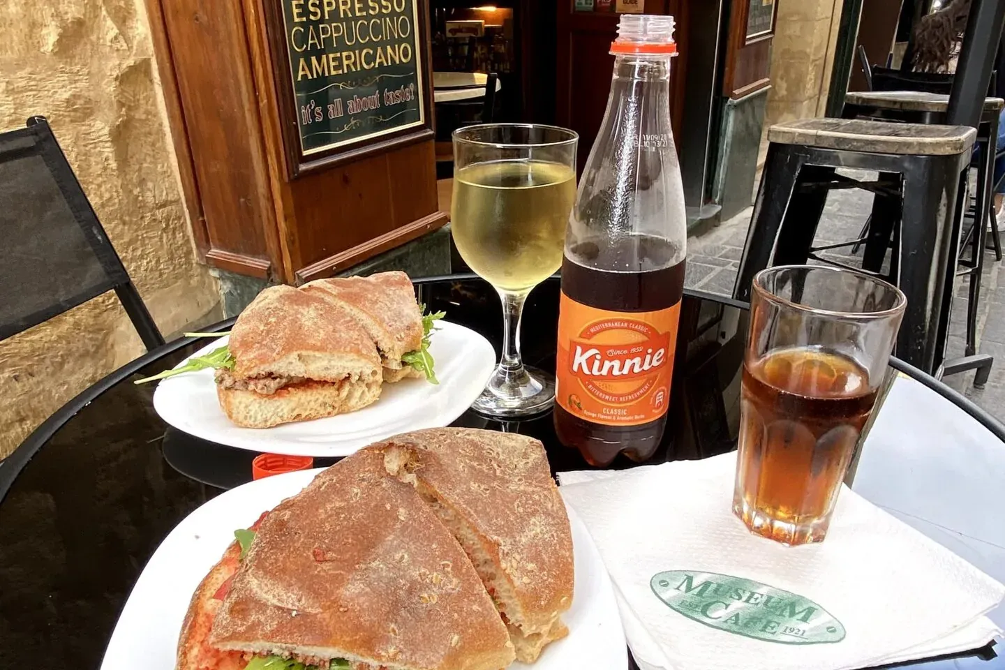 Mesa de café al aire libre con dos sándwiches de ciabatta, una copa de vino blanco, una botella de Kinnie y un refresco os...