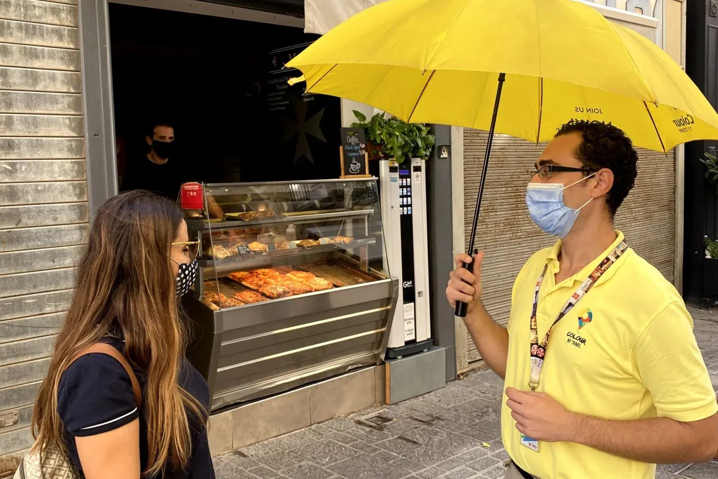 Hombre de camisa amarilla con paraguas habla con mujer frente a tienda 'Malta Pastizzi' con pasteles. Ambos con mascarillas.