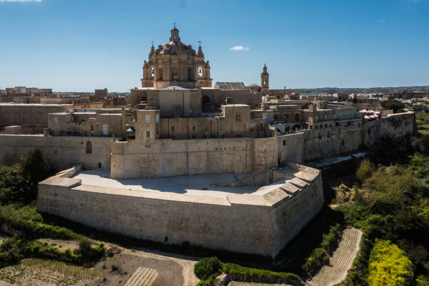 Vista aérea de una ciudad histórica fortificada con muros de piedra dorada, edificios y una gran catedral con cúpula bajo ...
