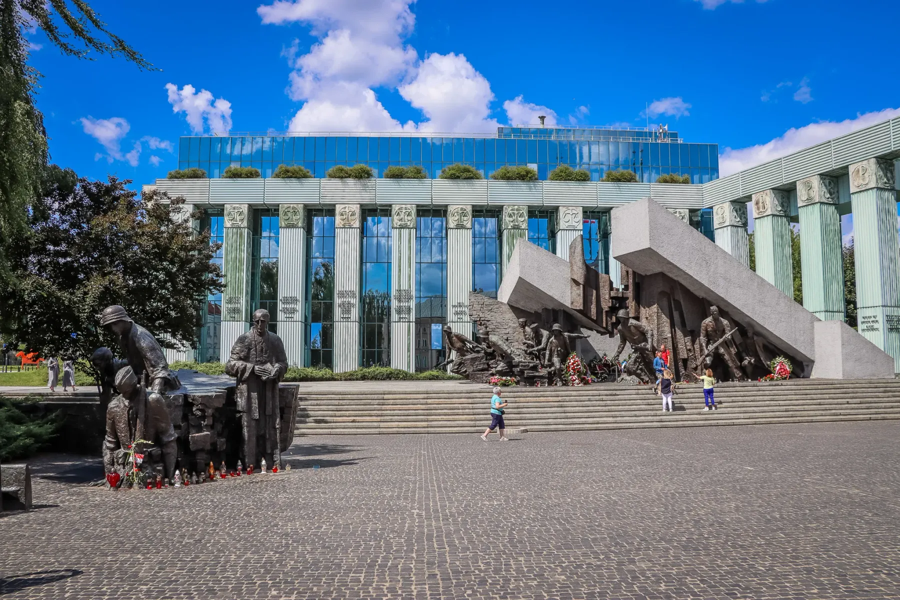 Brązowe posągi i duży betonowy pomnik na brukowanym placu w Warszawie. W tle nowoczesny budynek ze szklaną fasadą i kolumn...