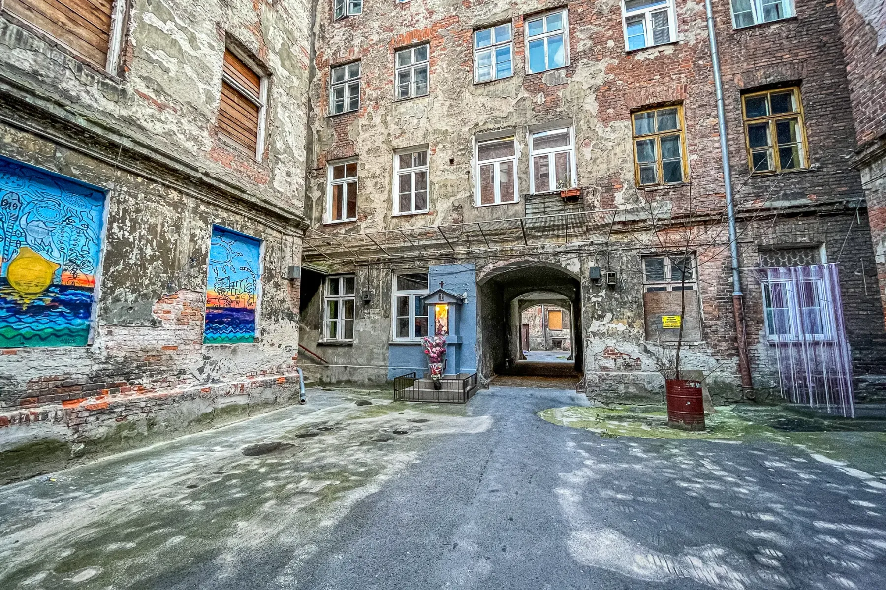Gritty tenement courtyard in Warsaw with peeling brick walls, a colorful street art mural, a small shrine, and an archway.