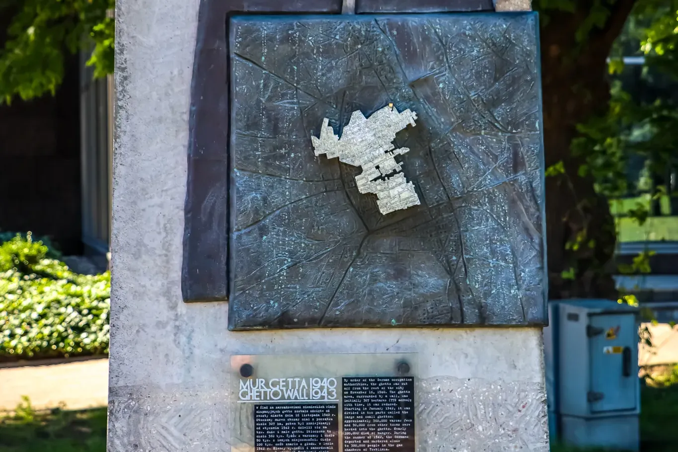 Concrete memorial pillar with a bronze plaque showing a map outline and text, 'Ghetto Wall 1940-1943', set in green surrou...
