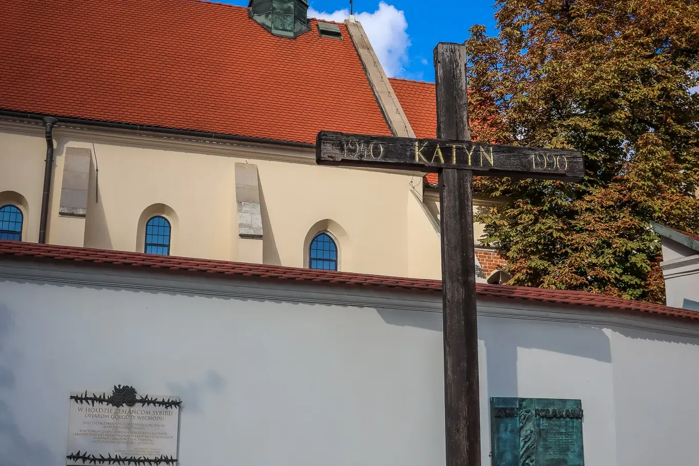 Großes hölzernes Katyn-Kreuz-Denkmal mit Inschrift 'KATYN 1940 1990', Gedenktafeln an weißer Mauer, Kirchendach und Turmsp...