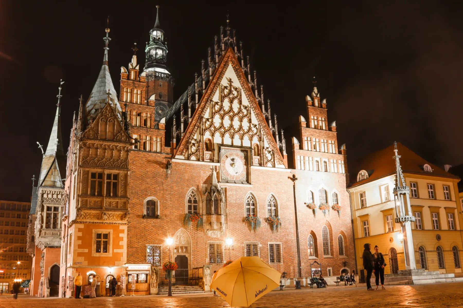 Podświetlony Ratusz we Wrocławiu nocą, z ozdobną fasadą, zegarem i wysoką wieżą na tle ciemnego nieba. Na placu żółty para...