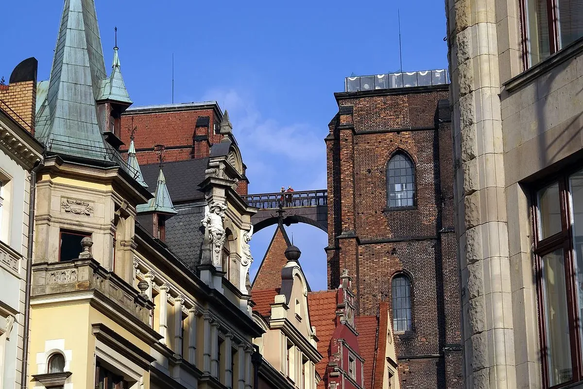 Widok na Most Pokutnic łączący dwie ceglane wieże, sfotografowany z ulicy pomiędzy historycznymi budynkami ze szpiczastymi...