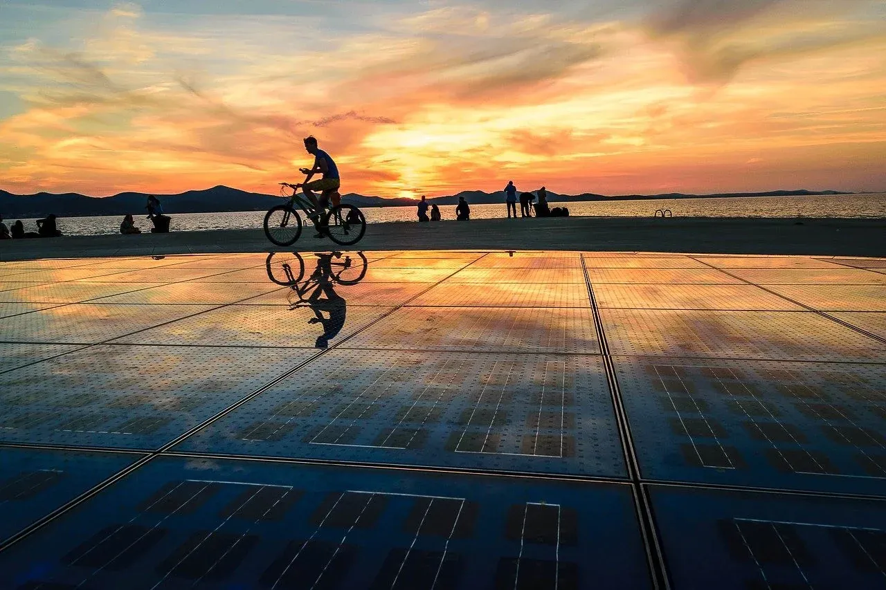 Persona en bicicleta silueteada contra una colorida puesta de sol sobre el mar, reflejada en una instalación solar.