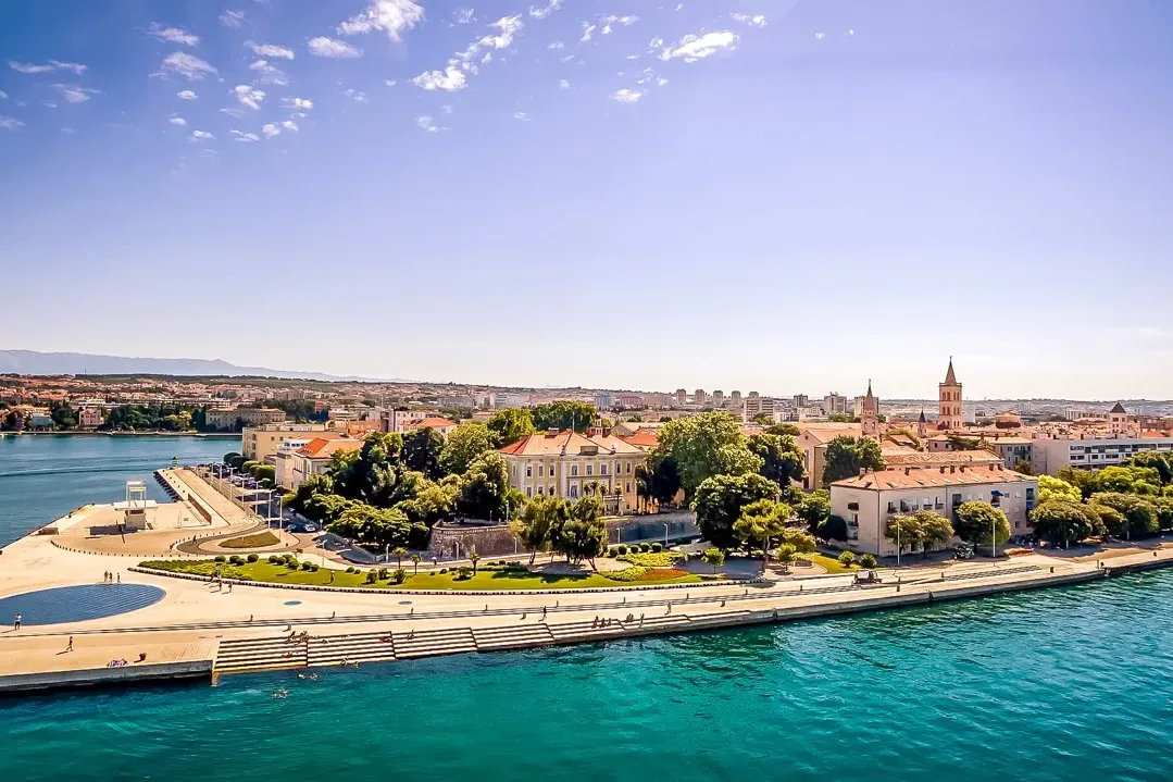 Vista aérea del paseo marítimo de Zadar con el Órgano Marino, edificios, mar azul claro y montañas lejanas.