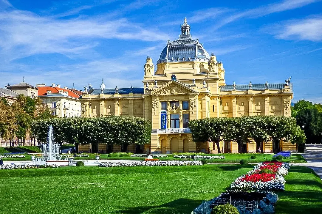 Pabellón de Arte neobarroco amarillo con cúpula de cristal, fuente en un parque verde con parterres y aguja de catedral le...