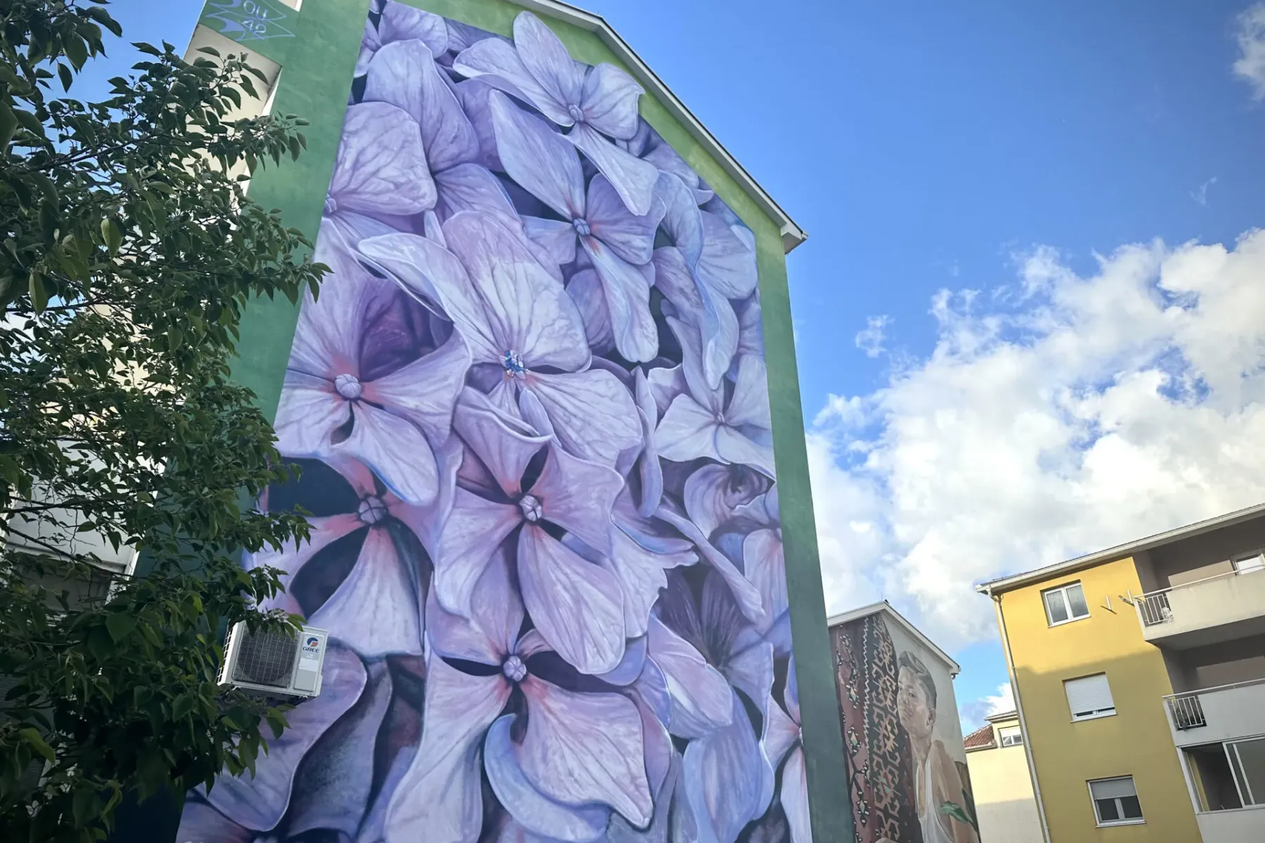 Duży mural fioletowych hortensji na zielonej fasadzie budynku. Widoczna gałąź drzewa i jednostka klimatyzacyjna.