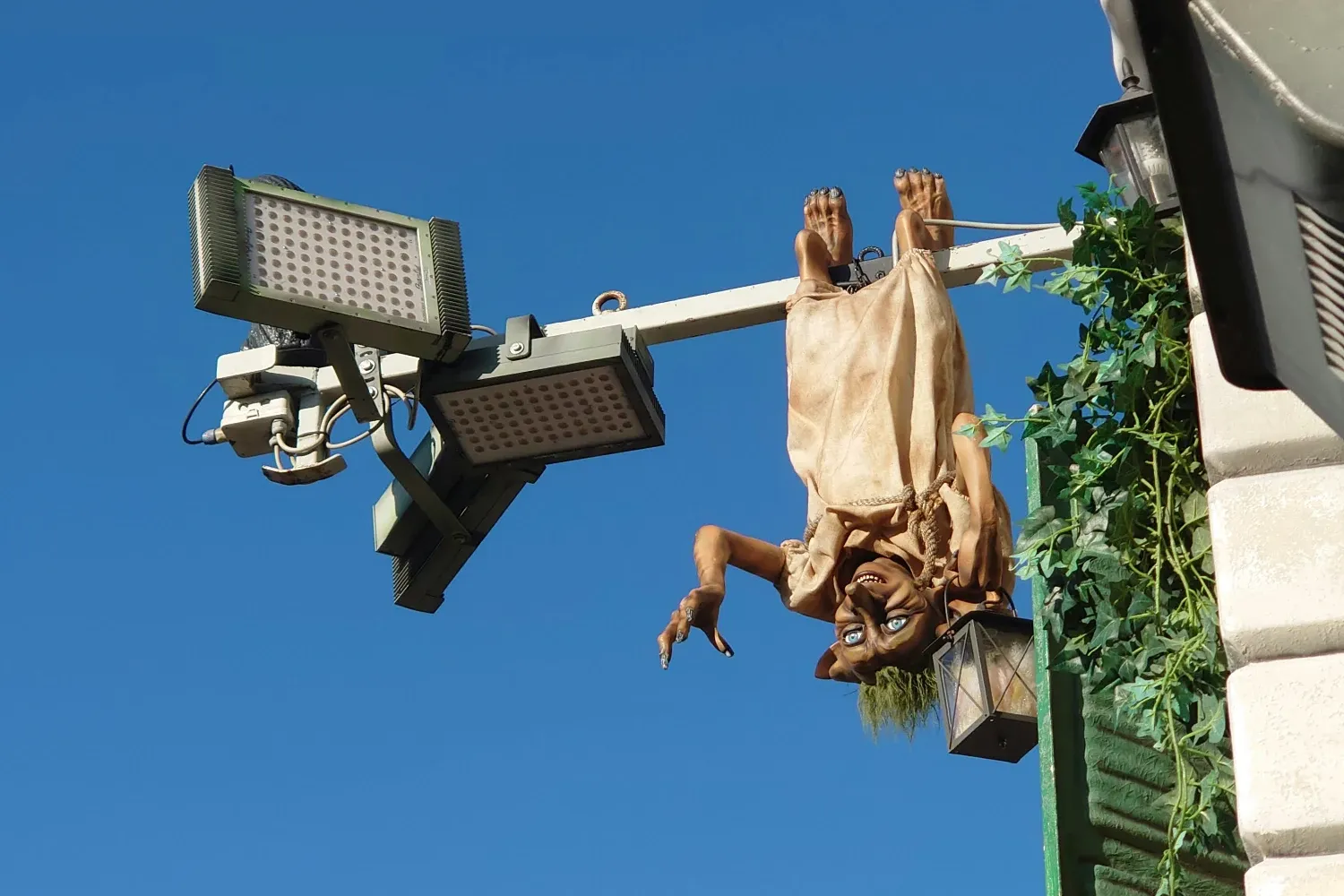 Una figura parecida a un duende cuelga boca abajo de un poste, sosteniendo un farol, con una noria al fondo.