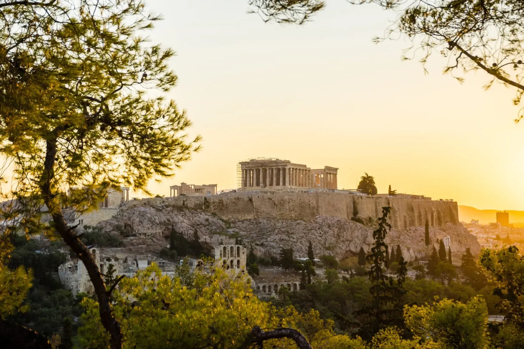 Die Akropolis von Athen mit Parthenon und Gerüsten, in warmes Sonnenuntergangslicht getaucht, durch grüne Bäume gesehen.