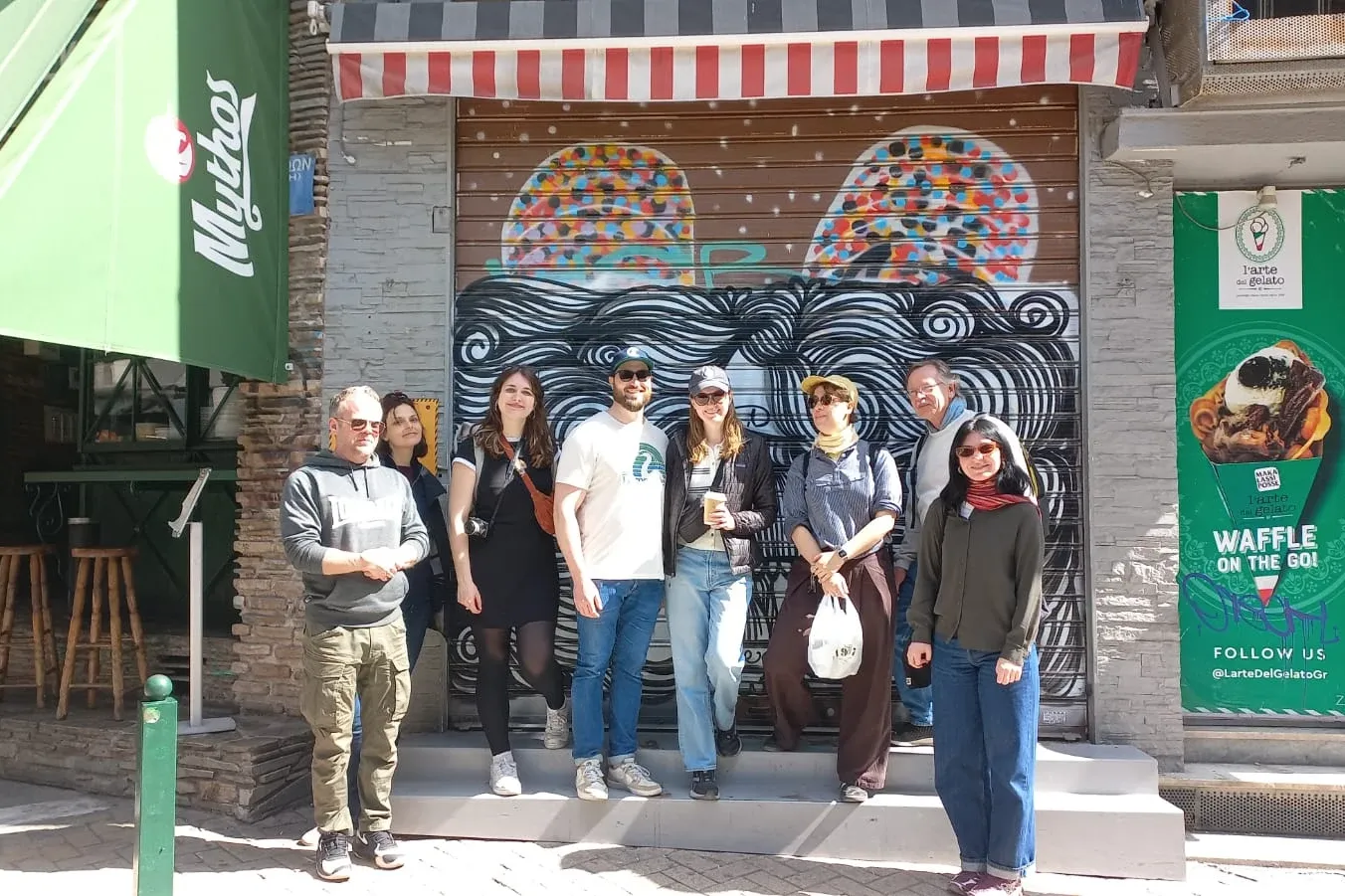 A group of tourists poses for a photo in front of a closed shop with graffiti on the streets of Athens.