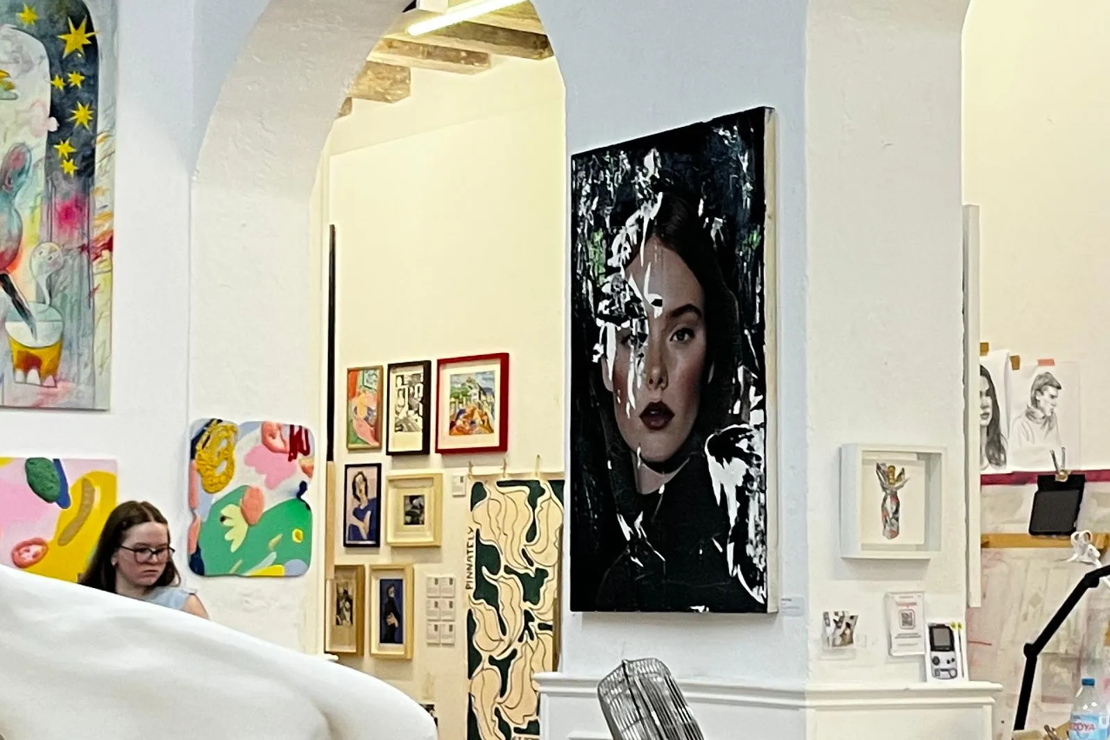 Art studio interior with paintings on white walls, exposed wooden beams, a partial white sculpture, and a woman observing ...