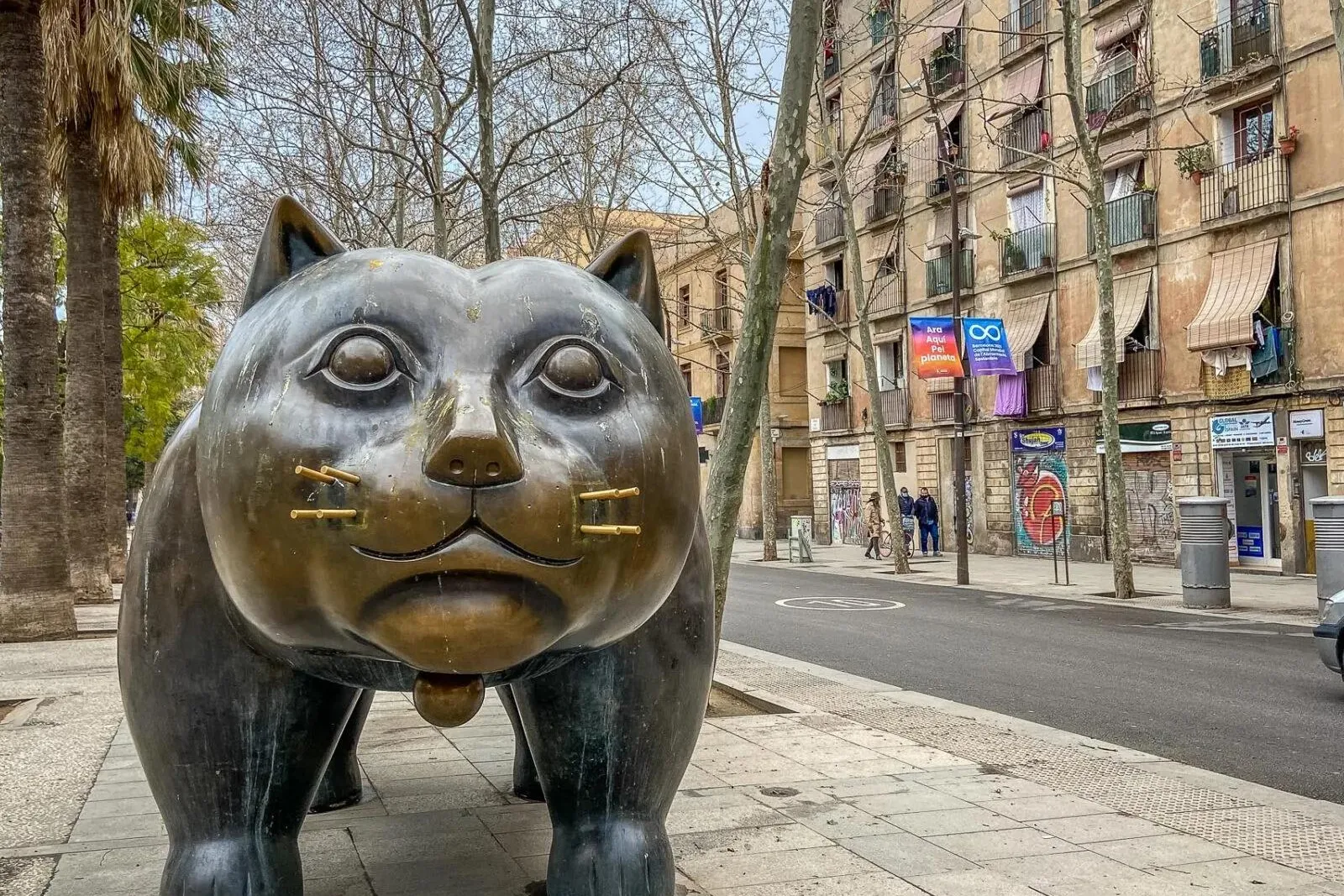 Large bronze sculpture of a plump cat with small ears, on a city sidewalk next to a street with buildings and trees.
