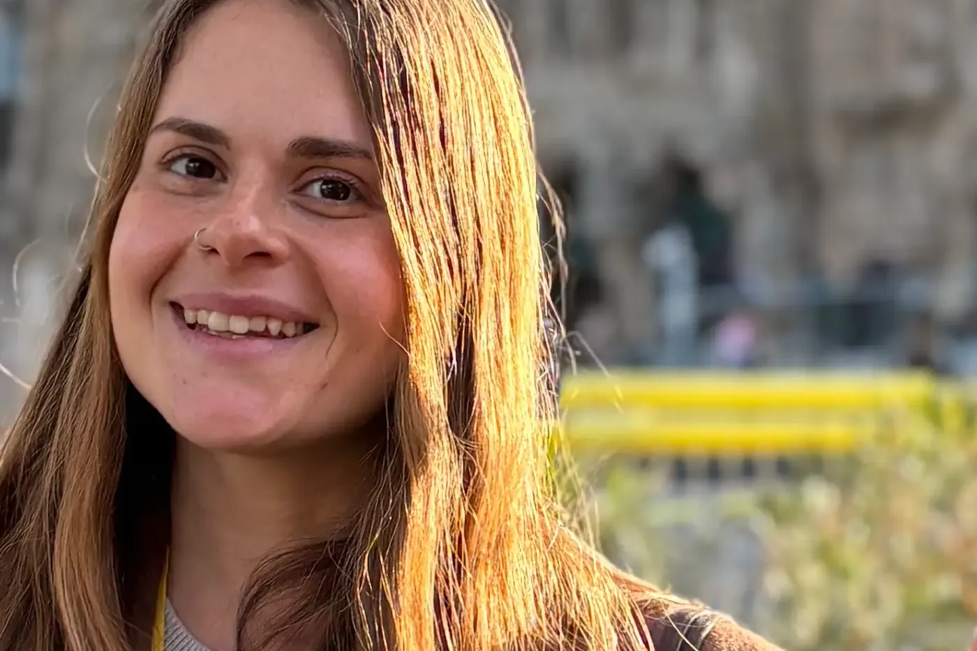 Mujer sonriente con abrigo marrón y cordón amarillo 'Guia de Turisme de Catalunya'. Edificio y palmas borrosos al fondo.