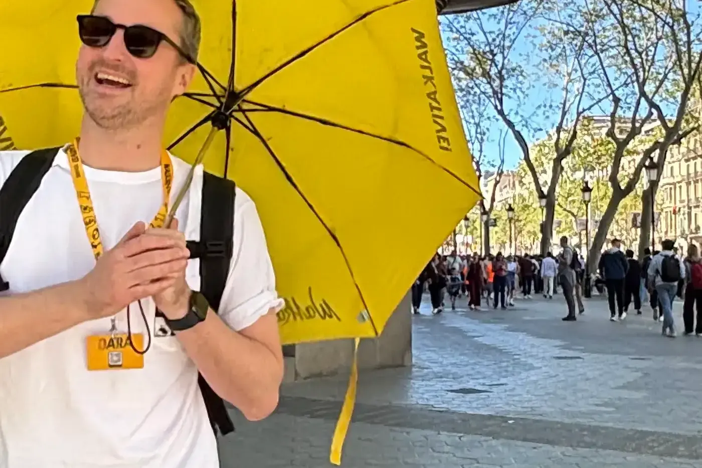 Guía turístico sonriente con paraguas amarillo de Walkative en una calle soleada de Barcelona. Edificio modernista ornamen...