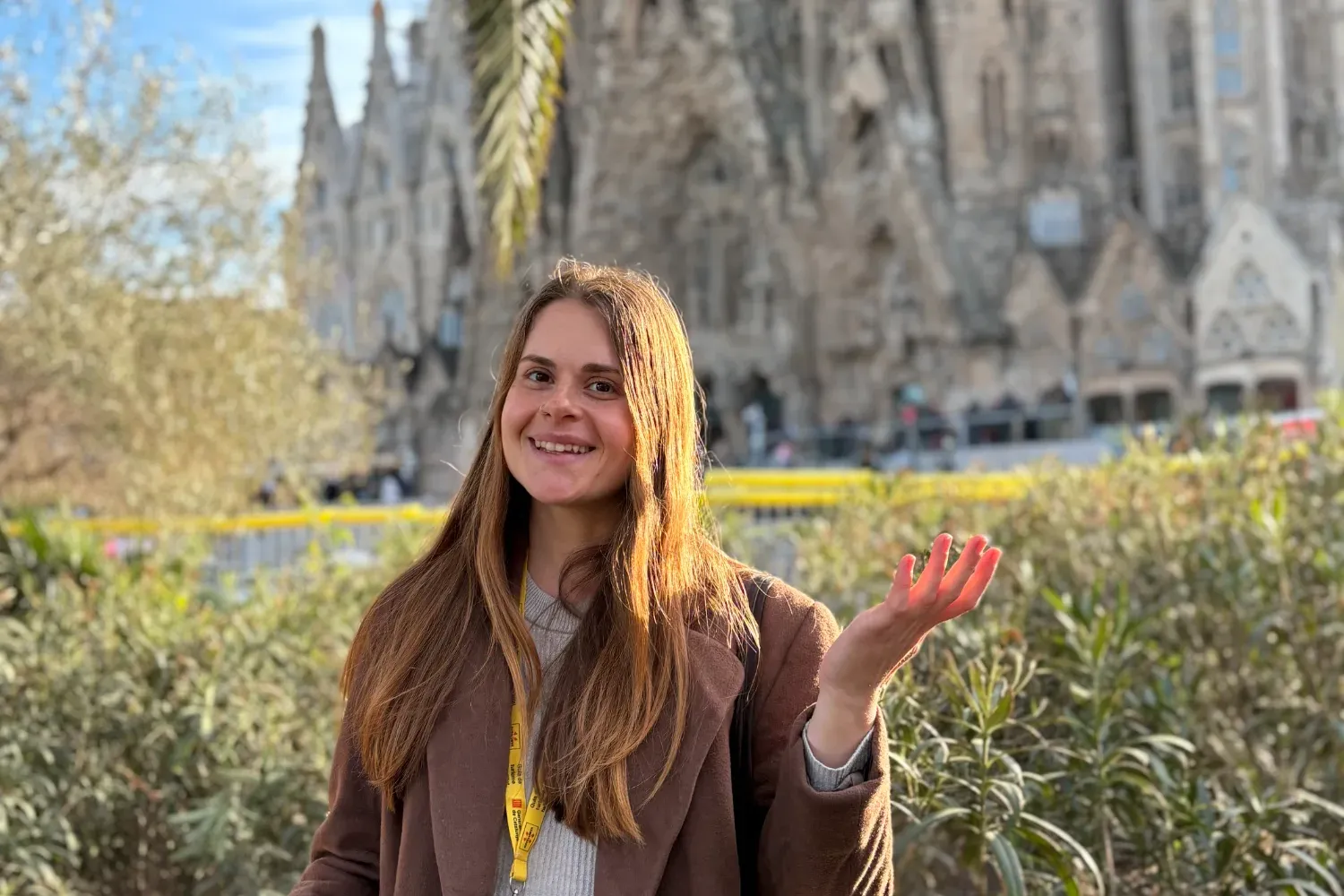 Uśmiechnięta przewodniczka Walkative w brązowym płaszczu przed bazyliką Sagrada Família Gaudiego w Barcelonie, obok palmy.