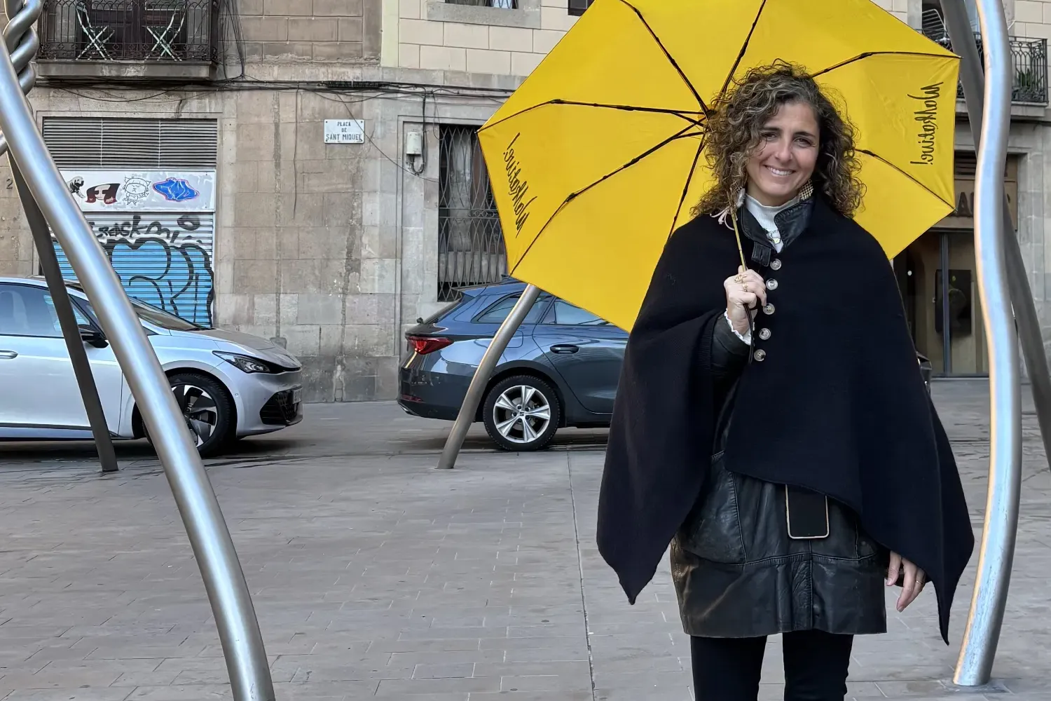 Mujer sonriente con pelo rizado sostiene un paraguas amarillo de Walkative en la Plaça de Sant Miquel, Barcelona. Edificio...