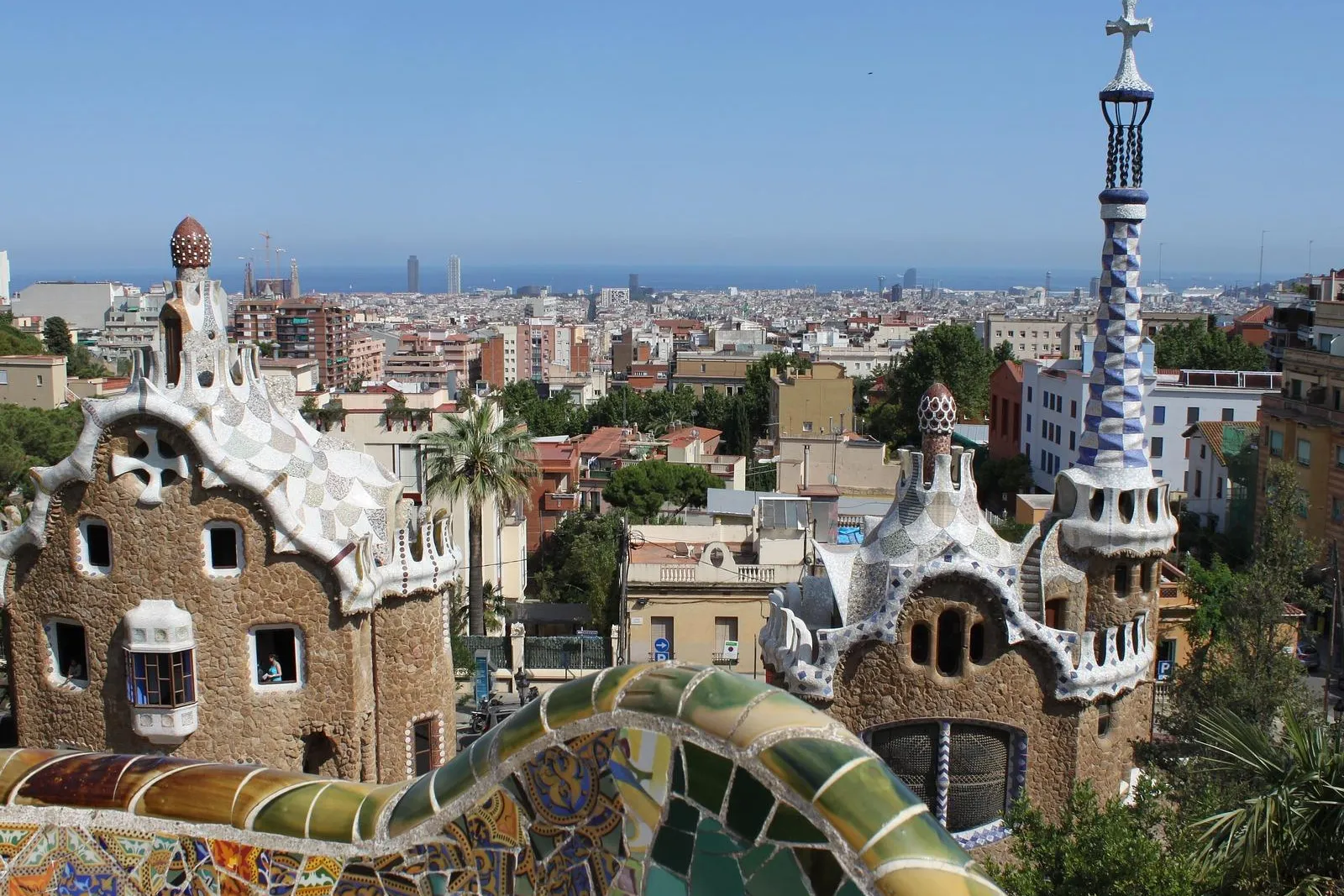 Blick vom Park Güell auf zwei Gaudi-Gebäude mit Mosaikdächern, eine bunte Fliesenmauer und Barcelonas Stadtbild mit Meer.